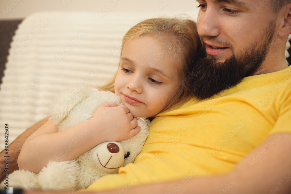 Beautiful little blonde girl leaning on her father's chest listeing what her father is reading. Bearded father embracing her little girl at home . Beautiful little blonde girl leaning on her father's chest listeing what her father is reading. Bearded father embracing her little girl at home .
