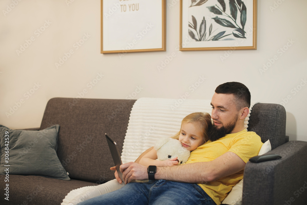Adorable little girl leaning on her father looking at a tablet at home on a couch. Lovely kid with her father spending time together. Adorable little girl leaning on her father looking at a tablet at home on a couch. Lovely kid with her father spending time together.