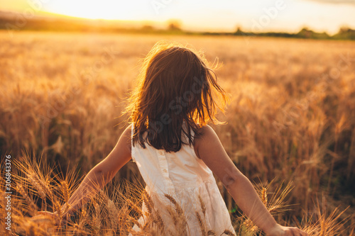 Back view of a lovely little girl walking in a wheat field touching wheat with hands against sunset.