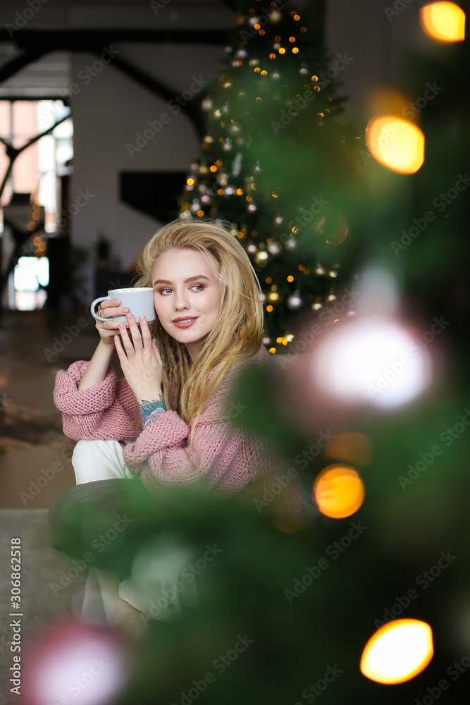 Portrait of a beautiful attractive happy bright young cute gently smiling blonde with dreadlocks in a pink cozy sweater with a Cup of coffee with marshmallows on the background of a Christmas tree. Portrait of a beautiful attractive happy bright young cute gently smiling blonde with dreadlocks in a pink cozy sweater with a Cup of coffee with marshmallows on the background of a Christmas tree.
