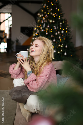 Portrait of a beautiful attractive happy bright young cute gently smiling blonde with dreadlocks in a pink cozy sweater with a Cup of coffee with marshmallows on the background of a Christmas tree.