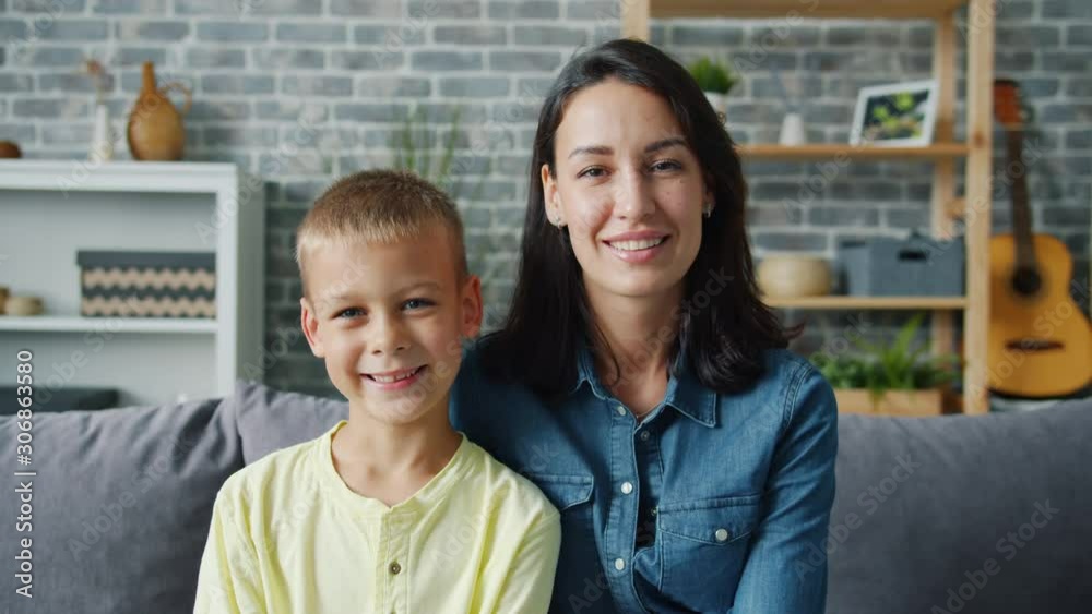Portrait of happy young family mother and little son smiling sitting together indoors looking at camera. Motherhood, childhood and apartment concept.