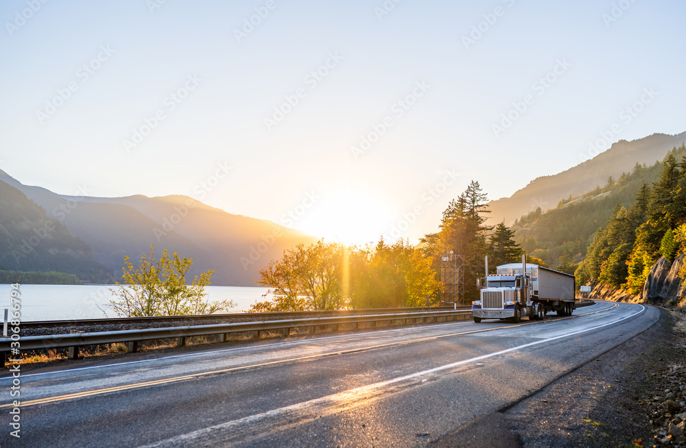 Big rig classic powerful semi truck transporting cargo in covered long bulk semi trailer running on the road along the river with mountain in sunset