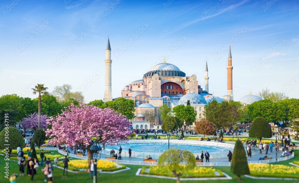 Naklejka premium Dzielnica Sultanahmet w Stambule w Turcji. Spacerujący ludzie, zielone pola trawiaste i fontanna w pobliżu słynnej świątyni Hagia Sophia. Dół zdjęcia jest zamazany.