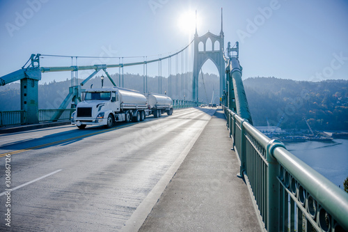 Big rig day cab white semi truck transporting liquid cargo in two tank semi trailers driving on the St Johns Bridge in Portland industrial area