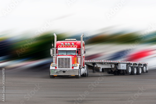 Red classic big rig semi truck with flat bed semi trailer turning on the parking lot on blurry background