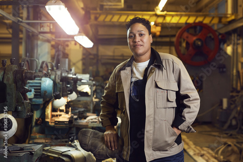 Waist up portrait of contemporary mixed-race woman posing in factory workshop at looking at camera, copy space