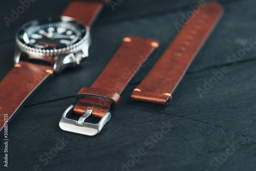 Close up of men's luxury watch with black bezel and handmade brown leather straps with steel buckles laying on the black wooden surface.