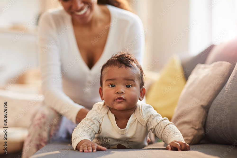 Warm toned portrait of cute African-American baby looking at camera while lying on couch with young mother doing massage, copy space Warm toned portrait of cute African-American baby looking at camera while lying on couch with young mother doing massage, copy space