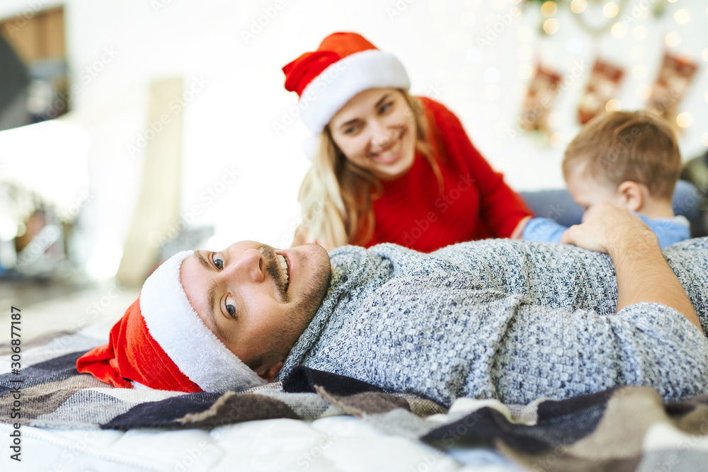 Portrait of handsome smiling man in red Santa cap, lying on the bed with little son and wife. Winter holiday, Christmas and New Year concept. happy time together. Portrait of handsome smiling man in red Santa cap, lying on the bed with little son and wife. Winter holiday, Christmas and New Year concept. happy time together.
