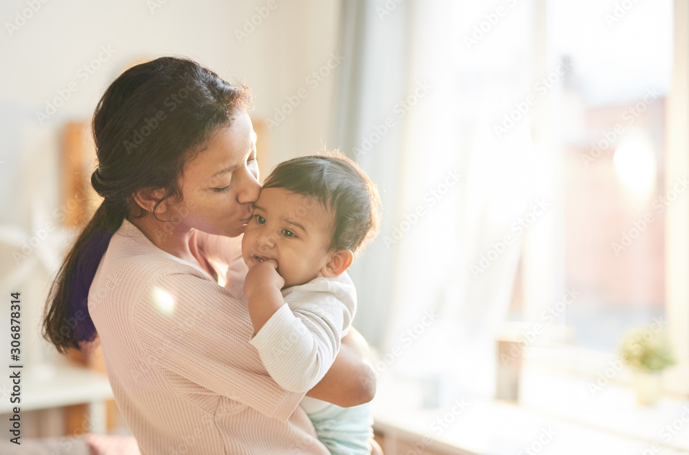 Young mother holding baby on her hands and kissing him while they are at home Young mother holding baby on her hands and kissing him while they are at home