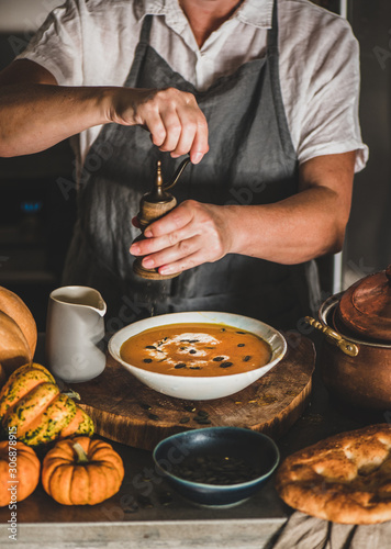 Eldery woman in grey linen apron cooking, tasting, putting pepper to Autumn seasonal pumpkin cream soup in modern kitchen interior. Fall seasonal comfort food
