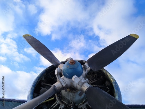 A four bladed black propeller on the nose of a green biplane aircraft against a blue sky