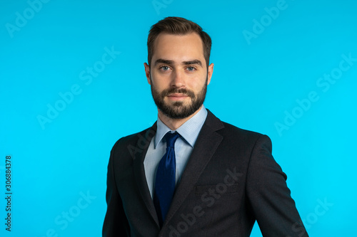 Close up portrait of young successful confident businessman with beard isolated on blue studio background. Man in business suit looking to camera and smiling. Portraiture of handsome guy.