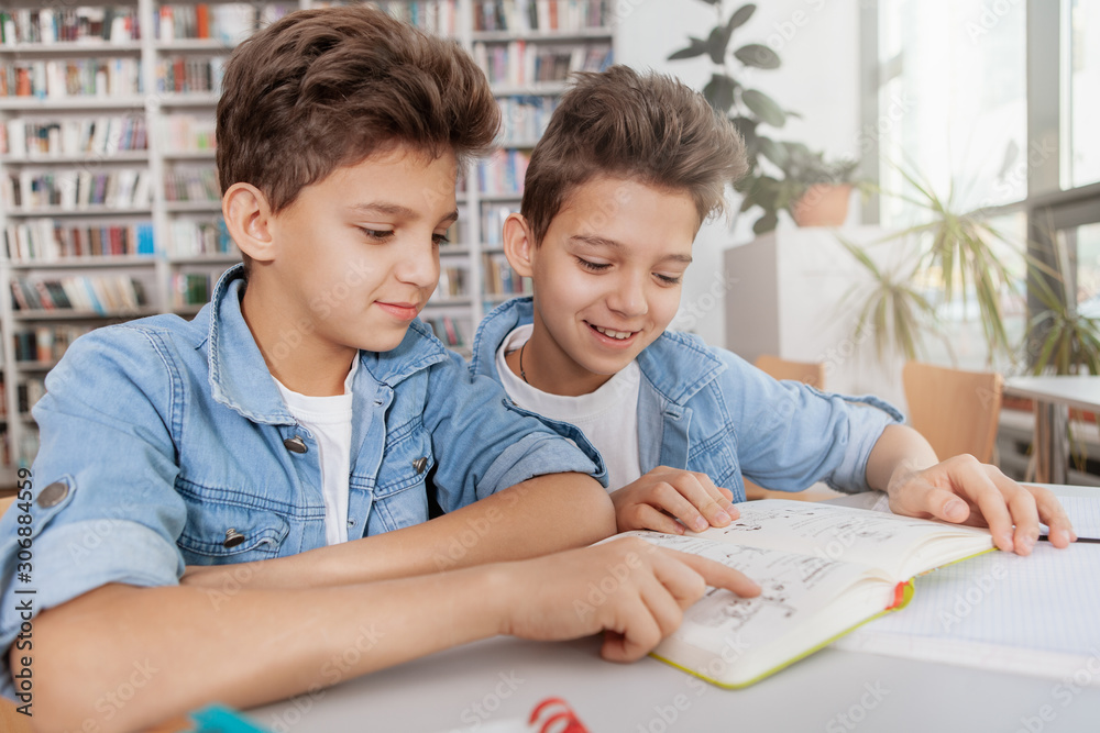 Two charming twin boys reading a book together at the library. School, education, friendship concept Two charming twin boys reading a book together at the library. School, education, friendship concept