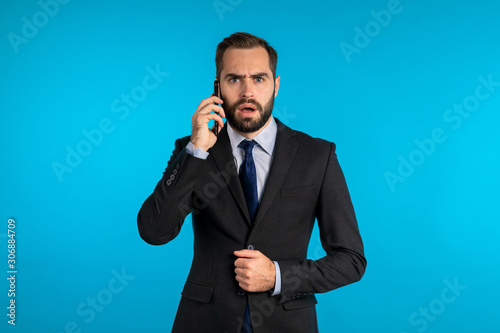 Businessman speaks on phone, heard bad news and expresses his amazement, shock isolated on blue studio background. Portrait of handsome man.