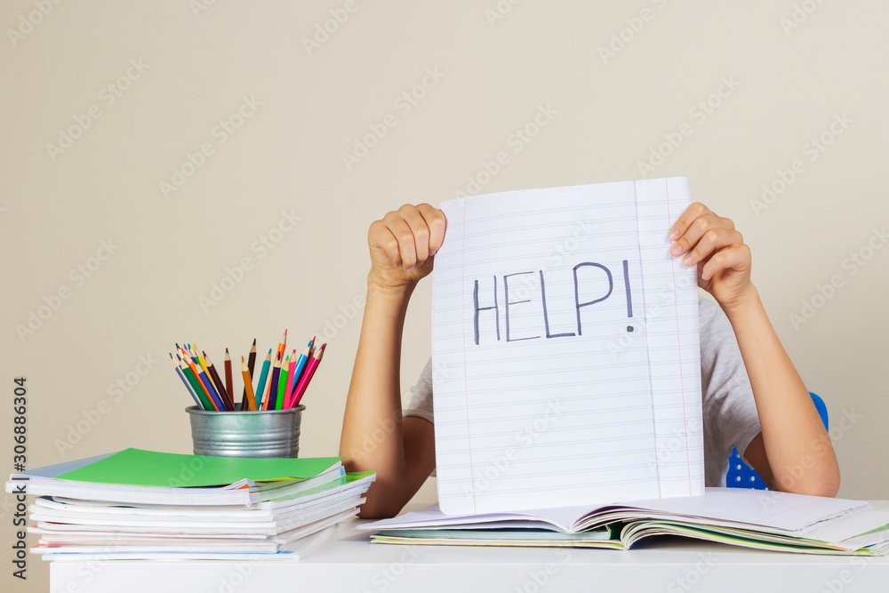 Help sign. Kid holding notebook with written word HELP. Kid sitting doing homework with pile of school books on the table Help sign. Kid holding notebook with written word HELP. Kid sitting doing homework with pile of school books on the table