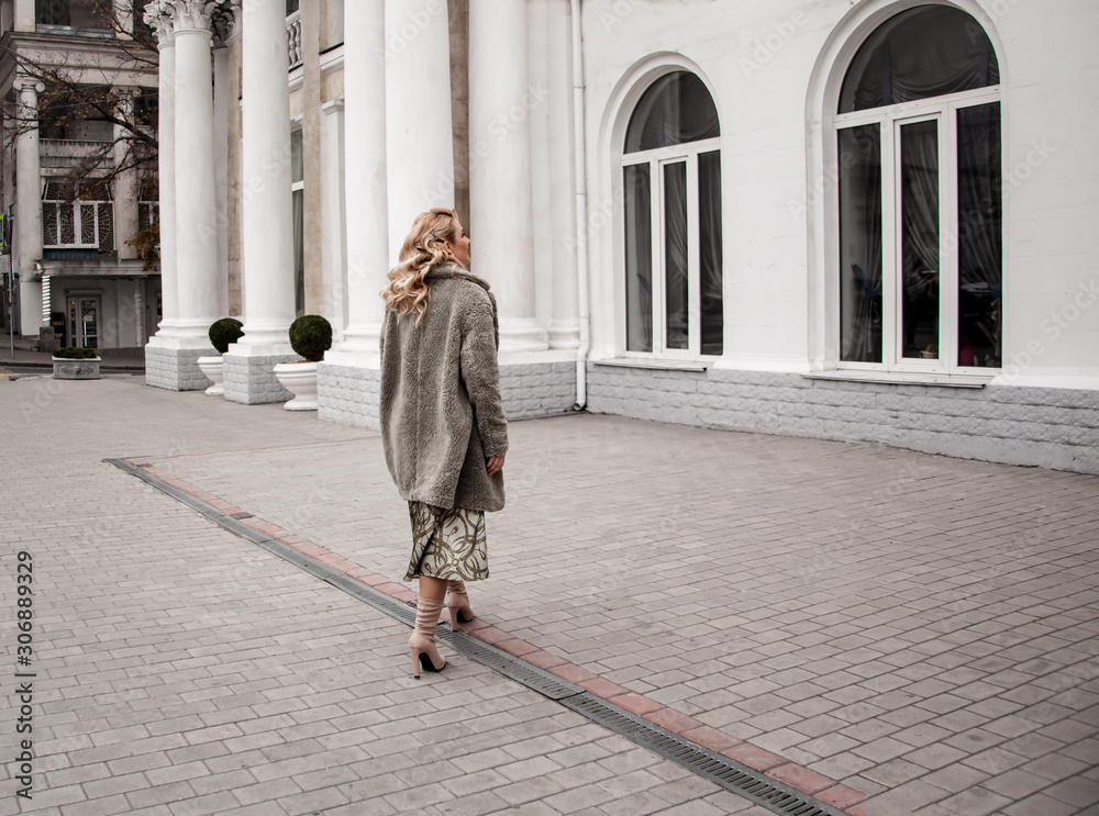 rear viev fashion european woman walking at the street, dressed modern clothes. Female moder wearing faux fur coat, tutleneck, skirt, and curl hairstyle