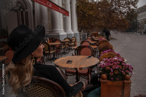 Beautiful 30s years old european woman sit in terrace of restourant and wiait the order. Female dressed black clothes and hat
