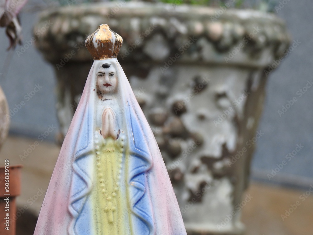 Scene in a cemetery: close-up of a small statue of Our Lady with a blue and pink cloak, a golden crown, clasped hands holding a rosary. In the blurred background, an old and worn stone vase.