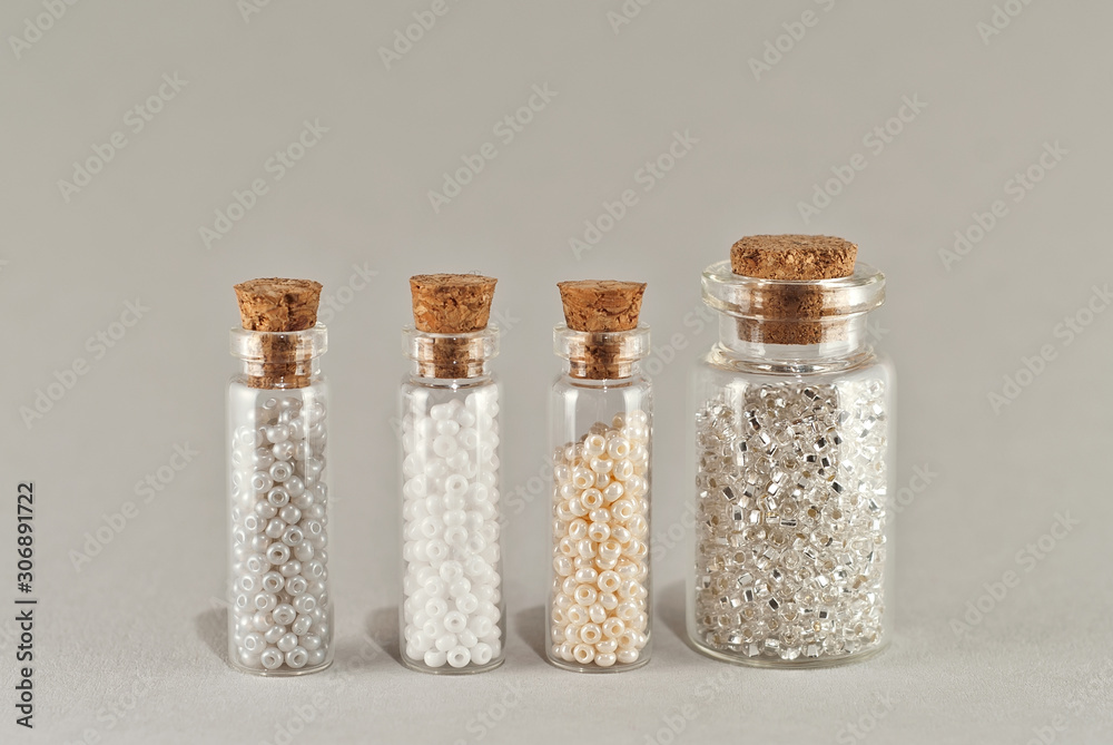 White, beige and yellow beads in glass jars on a gray background. Beads in a transparent container with a wooden cork. White, beige and yellow beads in glass jars on a gray background. Beads in a transparent container with a wooden cork.