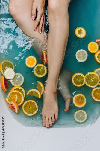 Woman's hand and legs in a bath filled with blue water and various cut citrus fruits