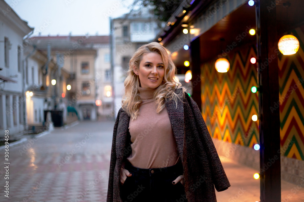 Fashion woman walking at the city, dressed modern clothes. Female model wearing coat, tutleneck, trousers and posing against the street with light at the evening