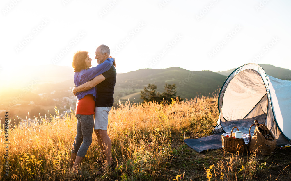 Senior tourist couple standing in nature at sunset, hugging. - obrazy, fototapety, plakaty 