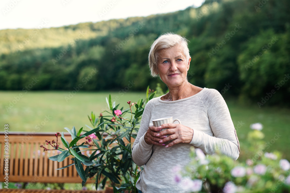 Senior woman with coffee standing outdoors on a terrace in summer. - obrazy, fototapety, plakaty 