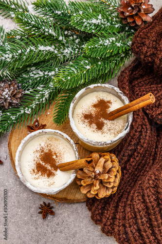 Homemade eggnog with cinnamon in glass. Typical Christmas dessert. Evergreen fir brunch, cones, cozy plaid, artificial snow. Stone concrete background