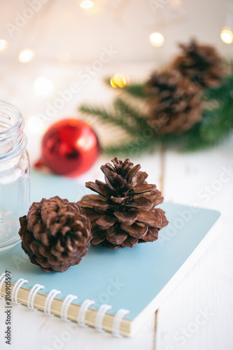 Pine cone or conifer cones on blue notebook near bottle and red bubble ball on white wood plank with golden light bokeh backdrop. Sweet vertical background for Christmas and winter season wallpaper.