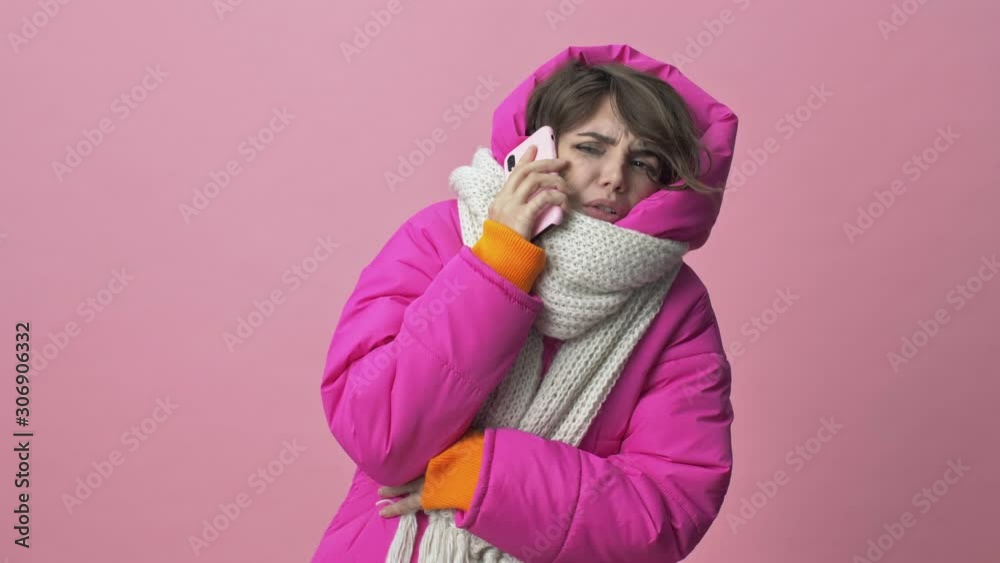 Frozen young woman wearing a winter jacket with a scarf is calling on the smartphone isolated over a pink background Frozen young woman wearing a winter jacket with a scarf is calling on the smartphone isolated over a pink background