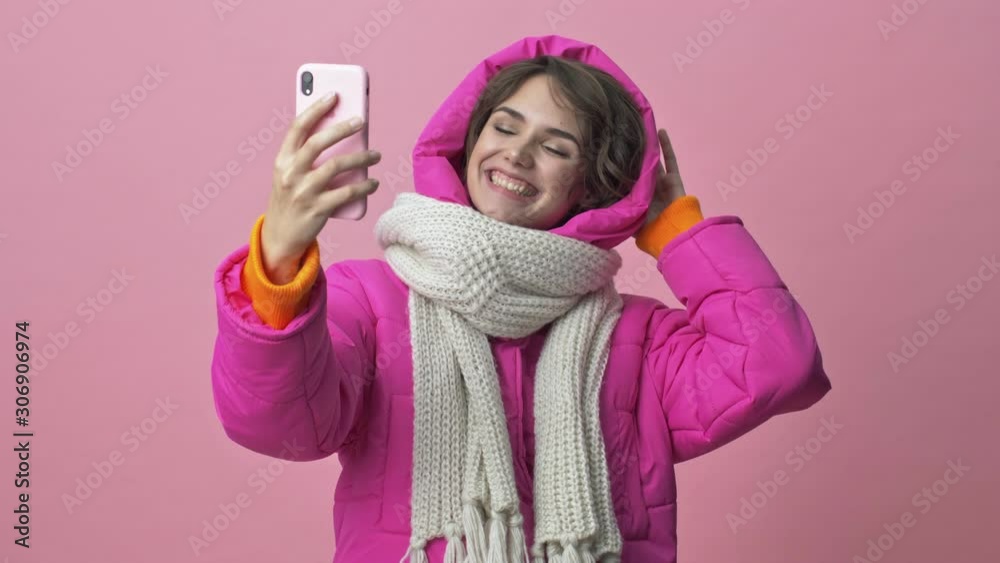 Joyful young woman wearing a winter jacket with a scarf is doing a selfie isolated over a pink background