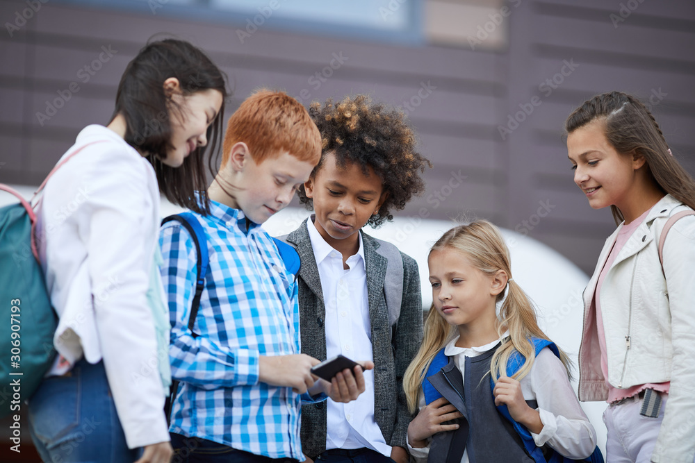 Group of classmates watching something on mobile phone together after school while standing outdoors Group of classmates watching something on mobile phone together after school while standing outdoors