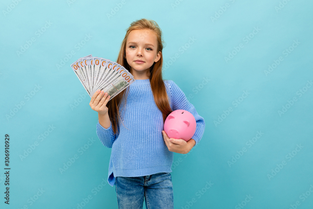 cute european girl holding a piggy bank and money in hands on a light blue  wall Photos | Adobe Stock