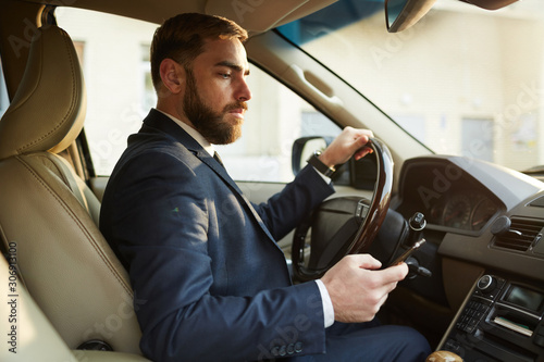 Young businessman sitting behind the steering wheel and driving while reading a message on his mobile phone