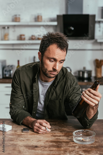 Cuadro en lienzo Upset man with alcohol depended holding cigarette and beer bottle at table