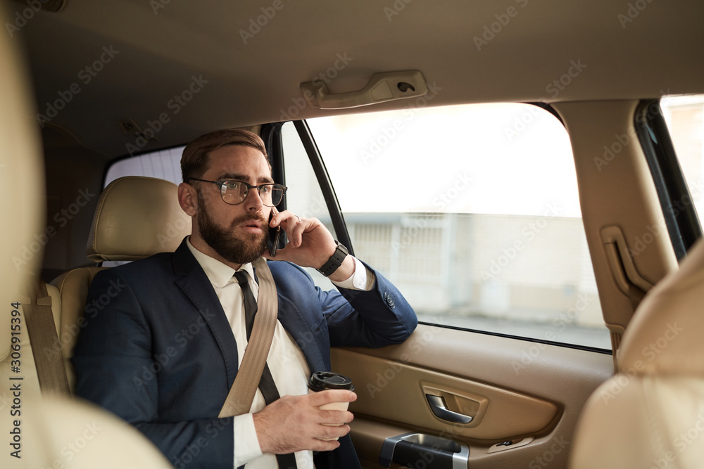 Worried young businessman talking on mobile phone and drinking coffee while sitting on back seat of a taxi