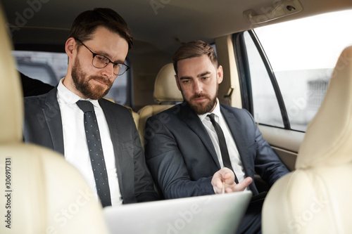 Two young businessmen sitting on back seat and discussing online presentation on laptop together during driving © AnnaStills