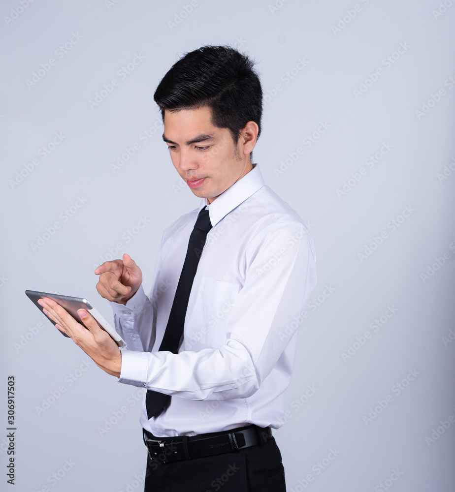 Portrait handsome young asian man wearing a white shirt holding smart phone or tablet smile and happy isolated on gray background in studio. Asian man people. business success concept. Portrait handsome young asian man wearing a white shirt holding smart phone or tablet smile and happy isolated on gray background in studio. Asian man people. business success concept.