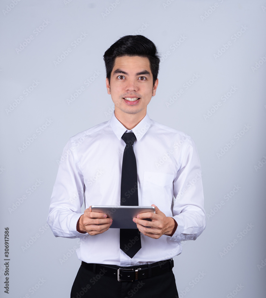 Portrait handsome young asian man wearing a white shirt holding smart phone or tablet smile and happy isolated on gray background in studio. Asian man people. business success concept. Portrait handsome young asian man wearing a white shirt holding smart phone or tablet smile and happy isolated on gray background in studio. Asian man people. business success concept.