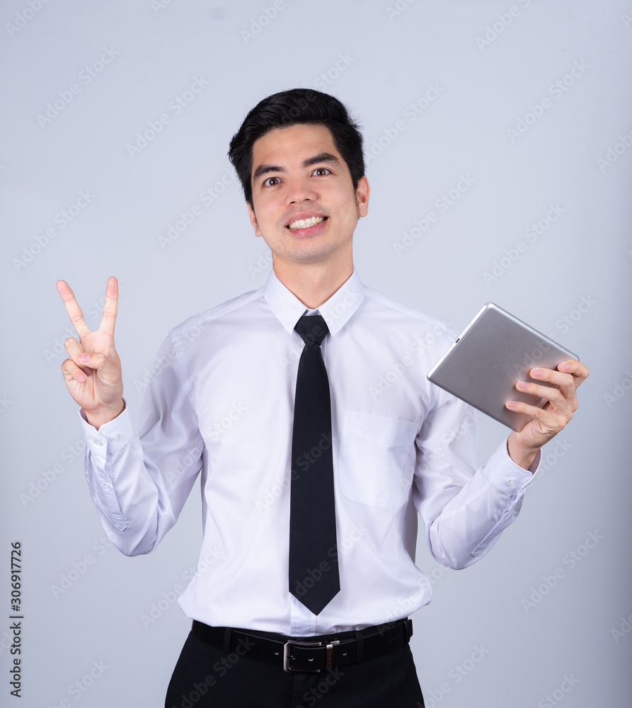 Portrait handsome young asian man wearing a white shirt holding smart phone or tablet smile and show two finger isolated on gray background in studio. Asian man people. business success concept. Portrait handsome young asian man wearing a white shirt holding smart phone or tablet smile and show two finger isolated on gray background in studio. Asian man people. business success concept.