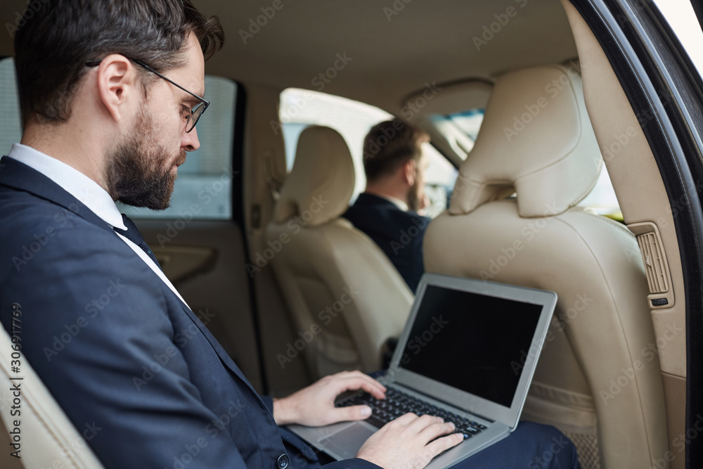 Young businessman in suit sitting on back seat of the car and working online using his laptop while driver driving the car