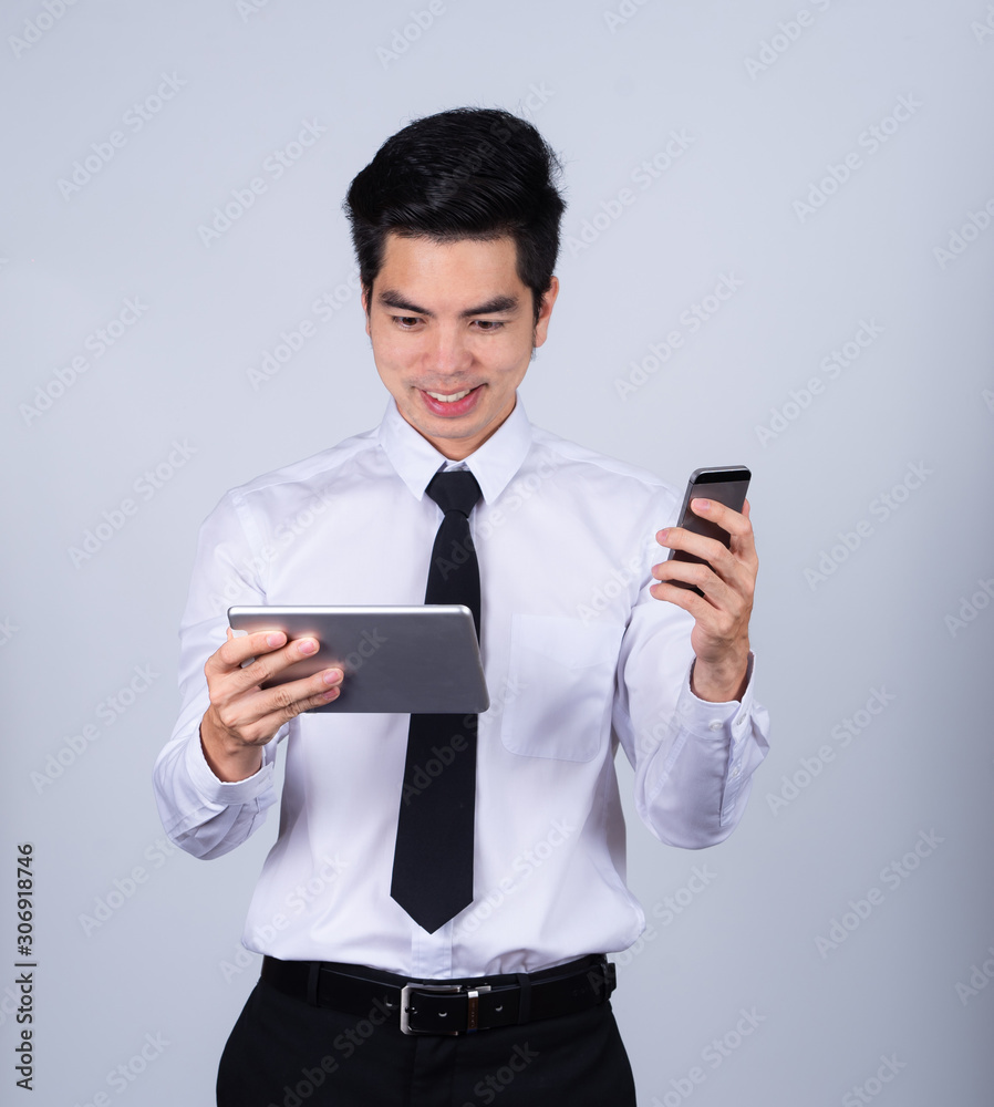 Portrait handsome young asian man wearing a white shirt holding smart phone or tablet smile and happy isolated on gray background in studio. Asian man people. business success concept. Portrait handsome young asian man wearing a white shirt holding smart phone or tablet smile and happy isolated on gray background in studio. Asian man people. business success concept.