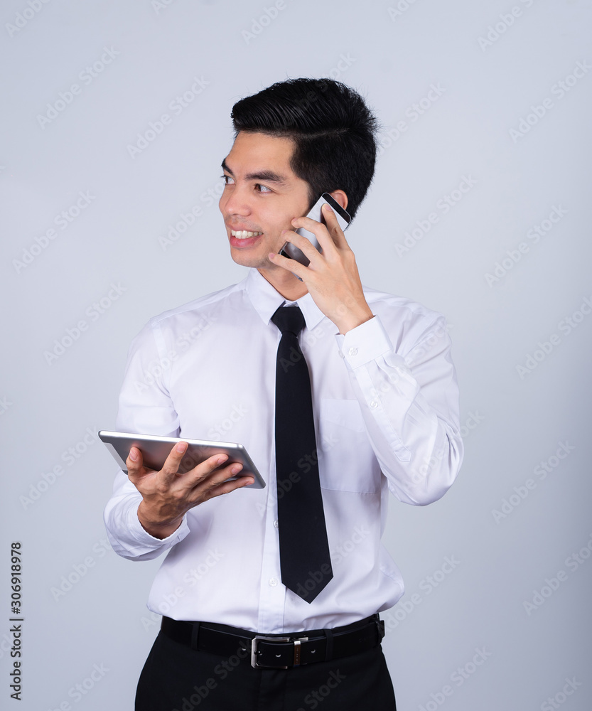 Portrait handsome young asian man wearing a white shirt takling telephone and holding tablet smile and happy isolated on gray background in studio. Asian man people. business success concept. Portrait handsome young asian man wearing a white shirt takling telephone and holding tablet smile and happy isolated on gray background in studio. Asian man people. business success concept.