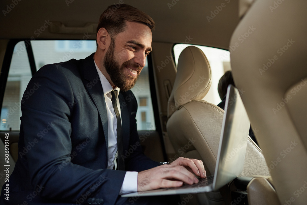 Young bearded businessman in suit sitting on back seat and typing on laptop computer during his journey in a taxi
