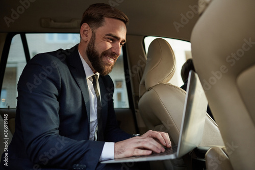 Young bearded businessman in suit sitting on back seat and typing on laptop computer during his journey in a taxi © AnnaStills
