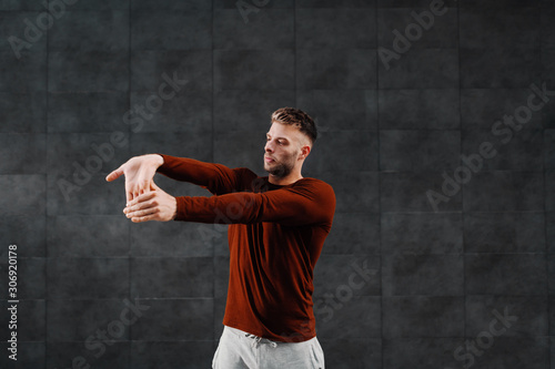 Handsome caucasian bearded sportsman in tracksuit doing warm up exercises for arms before running.