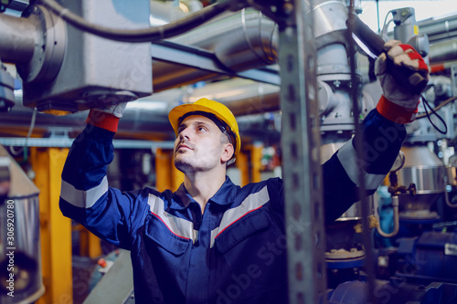 Handsome Caucasian blue collar worker in protective uniform and with hardhat on head checking on boiler while standing in factory. photo