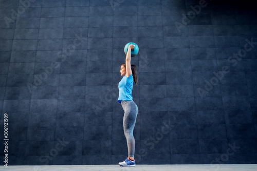 Side view of fit brunette in shape standing outdoors and lifting weight ball. In background is gray wall.